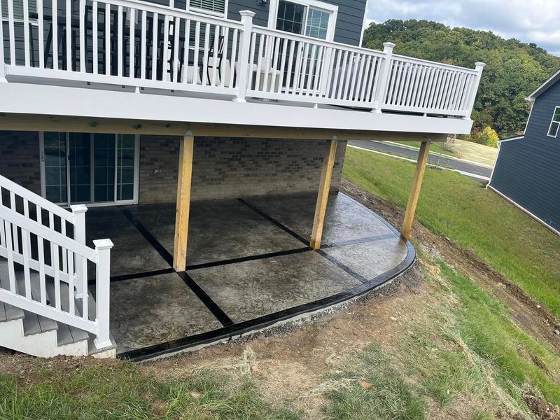 Backyard deck with concrete patio below, supported by wooden beams on a grassy hill.