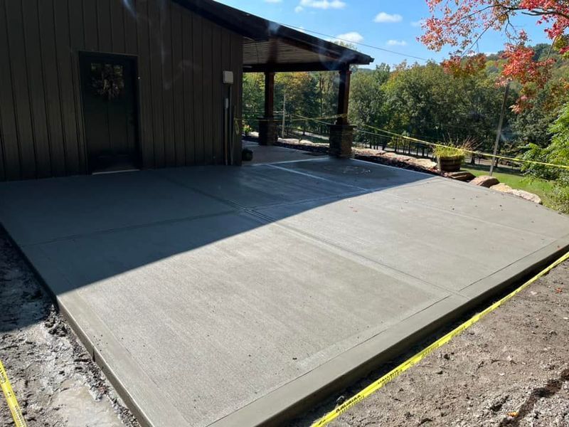Newly poured concrete patio adjacent to a wooden building, surrounded by dirt and grass.