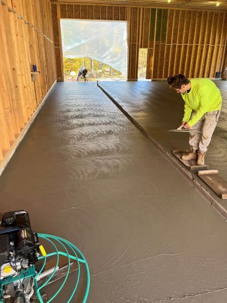 Workers smoothing wet concrete floor in a wooden-framed building with large opening.