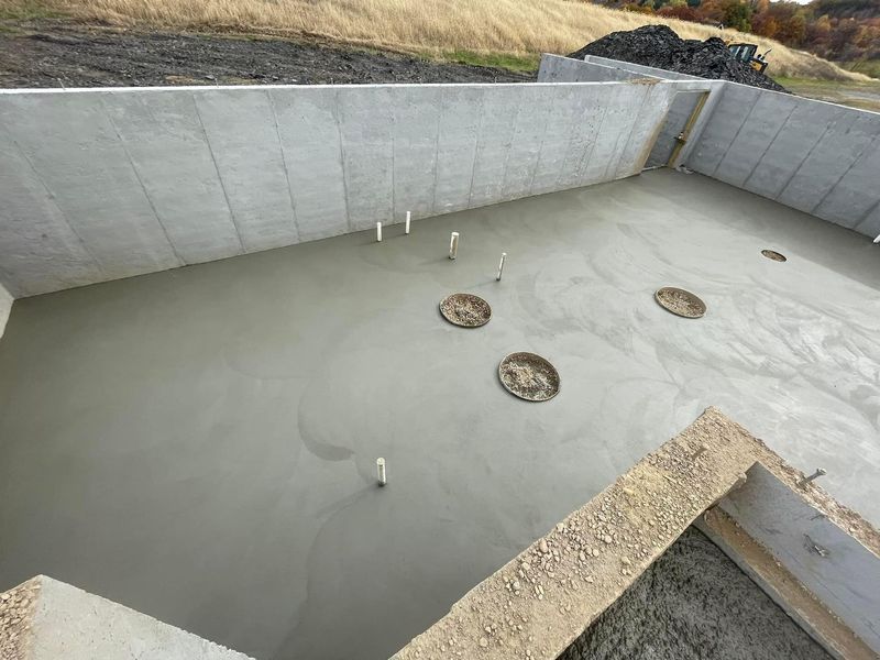 Newly poured concrete foundation with pipes and circular openings, surrounded by concrete walls.