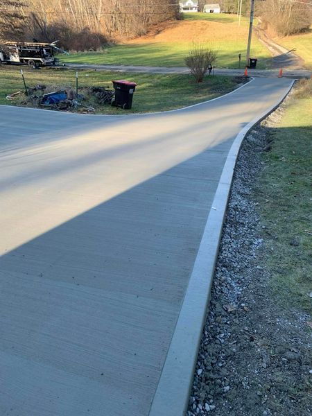 Newly poured concrete driveway curves toward a road, with grass on the right and a grassy area in the background.