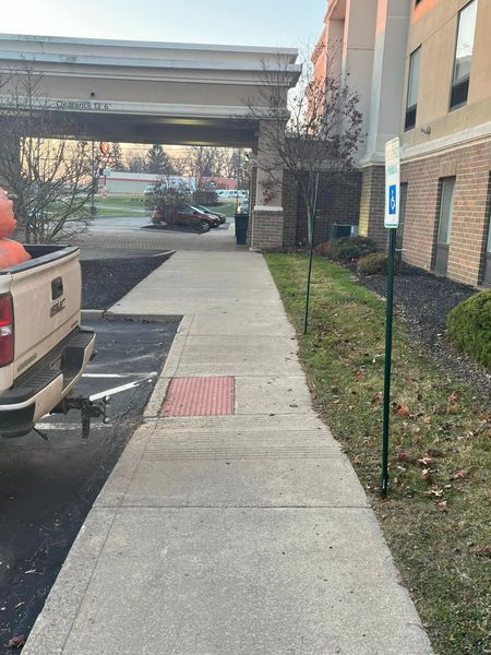 Sidewalk leading to hotel entrance with accessible parking sign, next to a truck and some bushes.