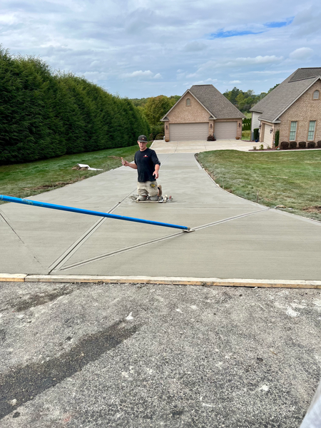 Man using a screed on a newly poured concrete driveway. Houses and trees in the background. Cloudy day.