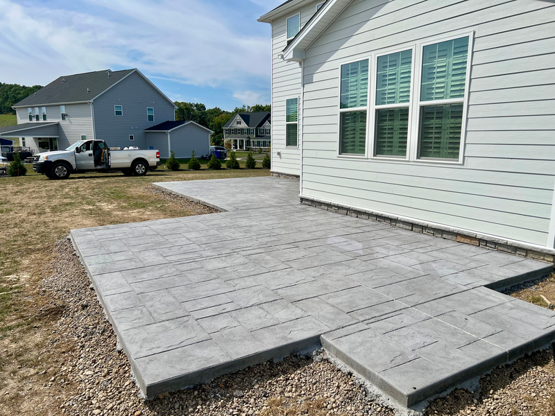 Gray concrete patio next to a white house, with a truck parked nearby in a grassy yard.