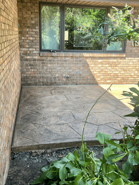 Concrete patio with brick wall, window above, and greenery to the right.