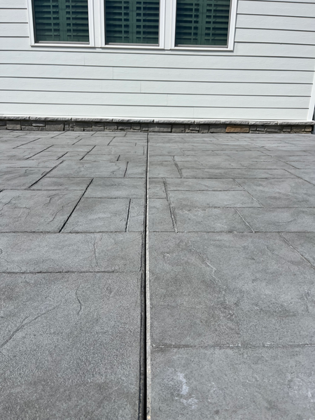 Gray stamped concrete patio with expansion joint against a white house with window.
