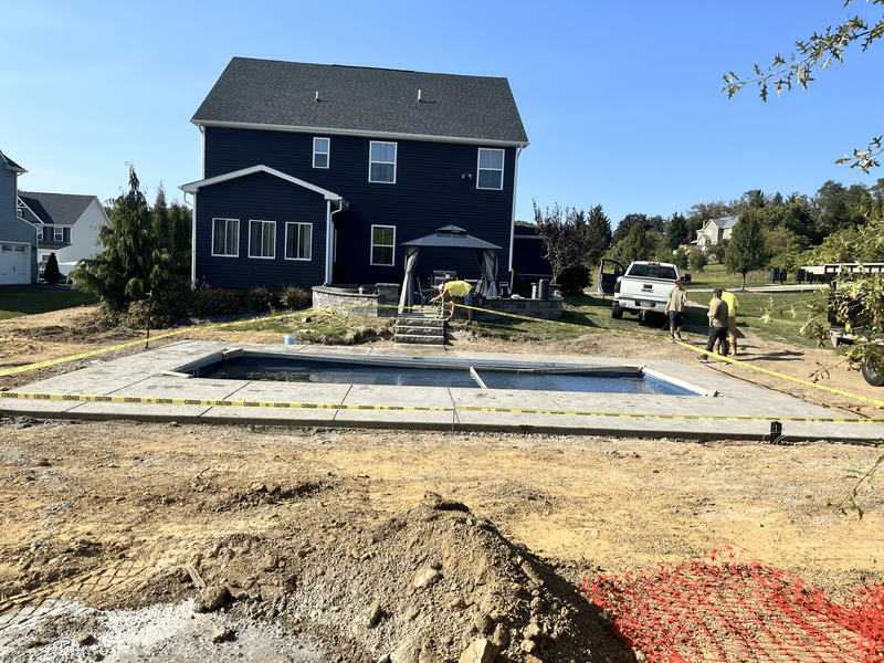 Pool under construction in backyard of blue house. Construction workers, concrete, dirt, and safety tape.