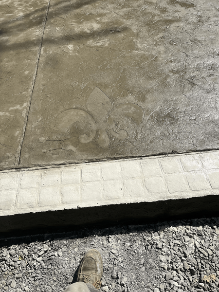 Concrete slab with a fleur-de-lis design, viewed from above, with a person's shoe and gravel visible below.