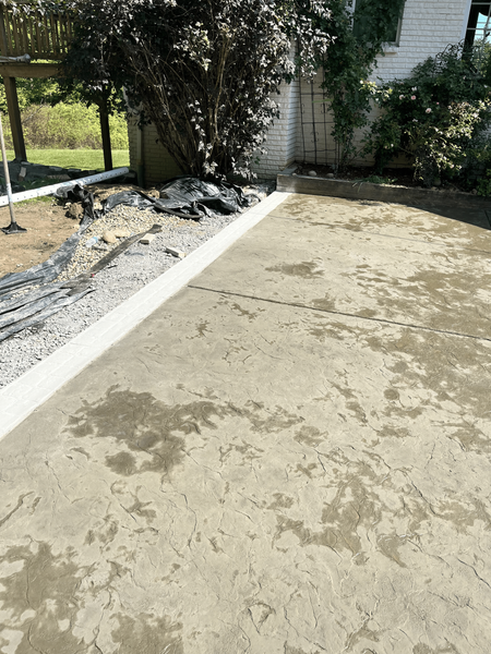 Newly poured concrete patio with a white border next to a house, bushes and a grassy area.