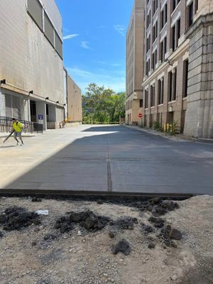Concrete alley between two buildings; a person uses a leaf blower. Bright sky in the distance.