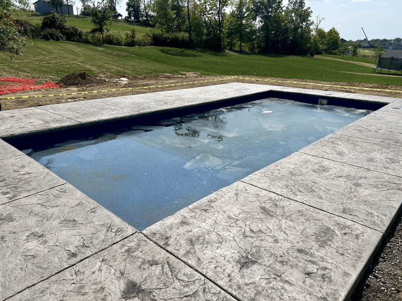 Rectangular in-ground pool with gray stamped concrete border surrounded by grassy hill under blue sky.