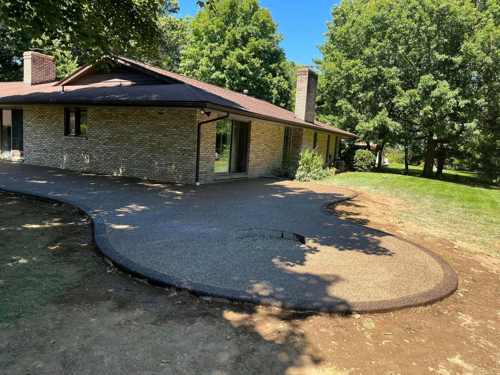 Stone house with a pebble patio, bordered by dark stone, grassy yard, trees, and blue sky.