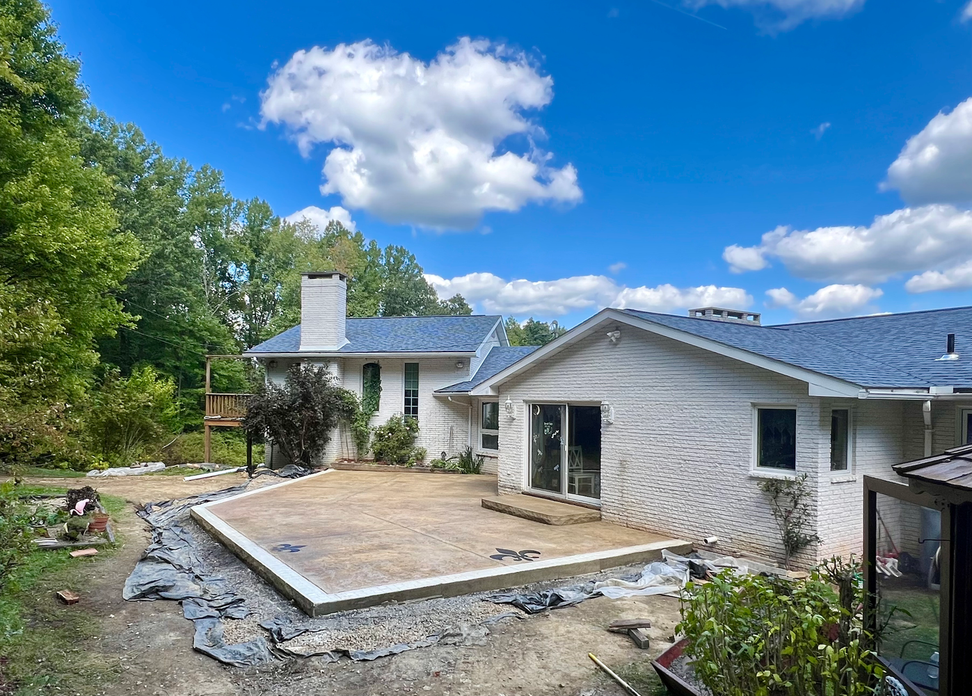 Backyard patio construction with house, blue sky, trees. Gray concrete foundation, dirt ground.