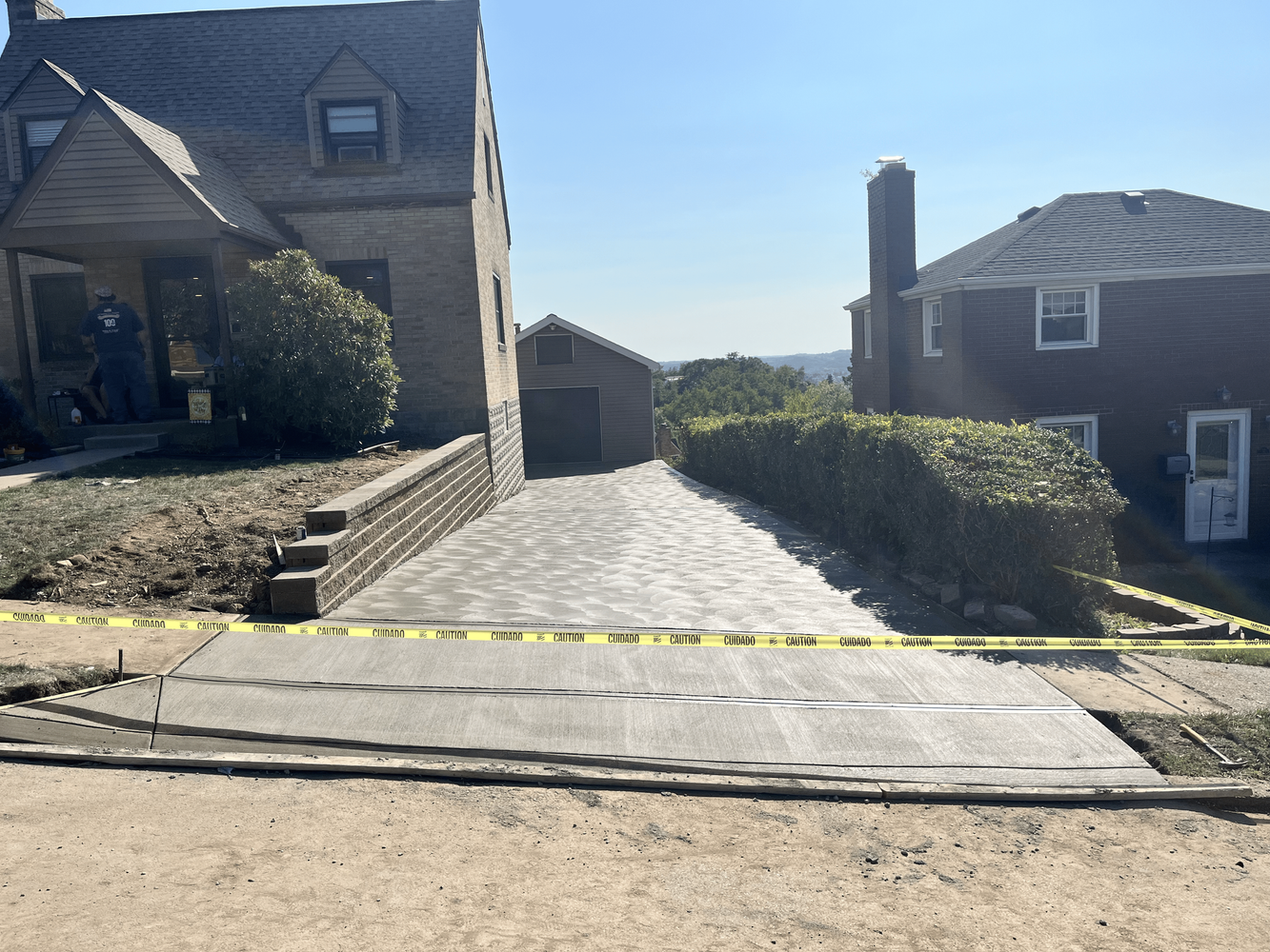 Driveway with fresh concrete, yellow caution tape, houses on either side, sunny day.