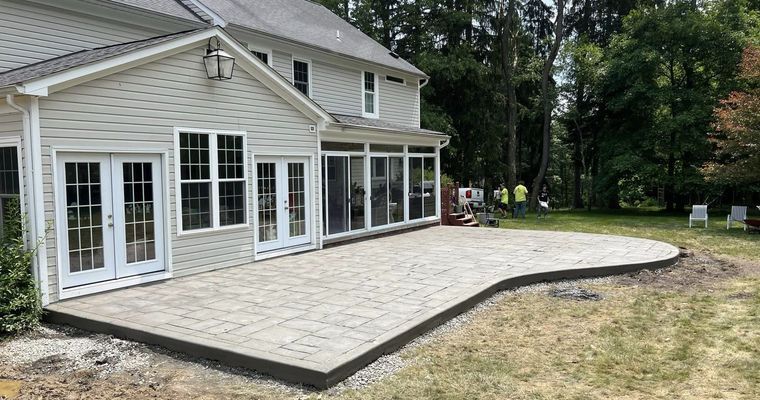 Backyard patio with a house. Gray pavers and trim. Green grass, trees, and blue sky.