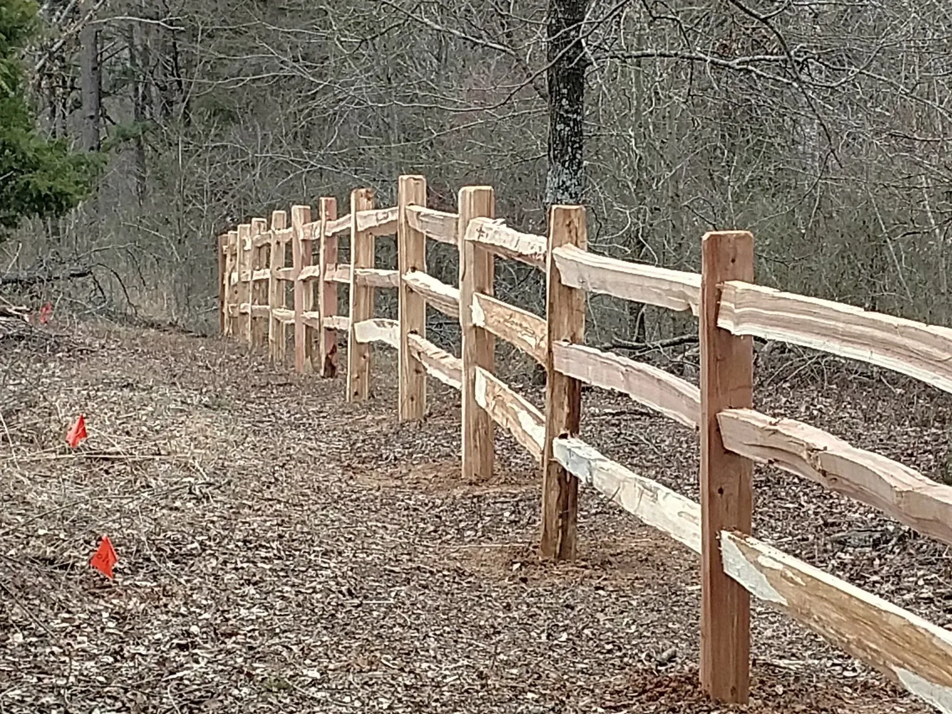Wooden split-rail fence in a wooded area, with fallen leaves on the ground.