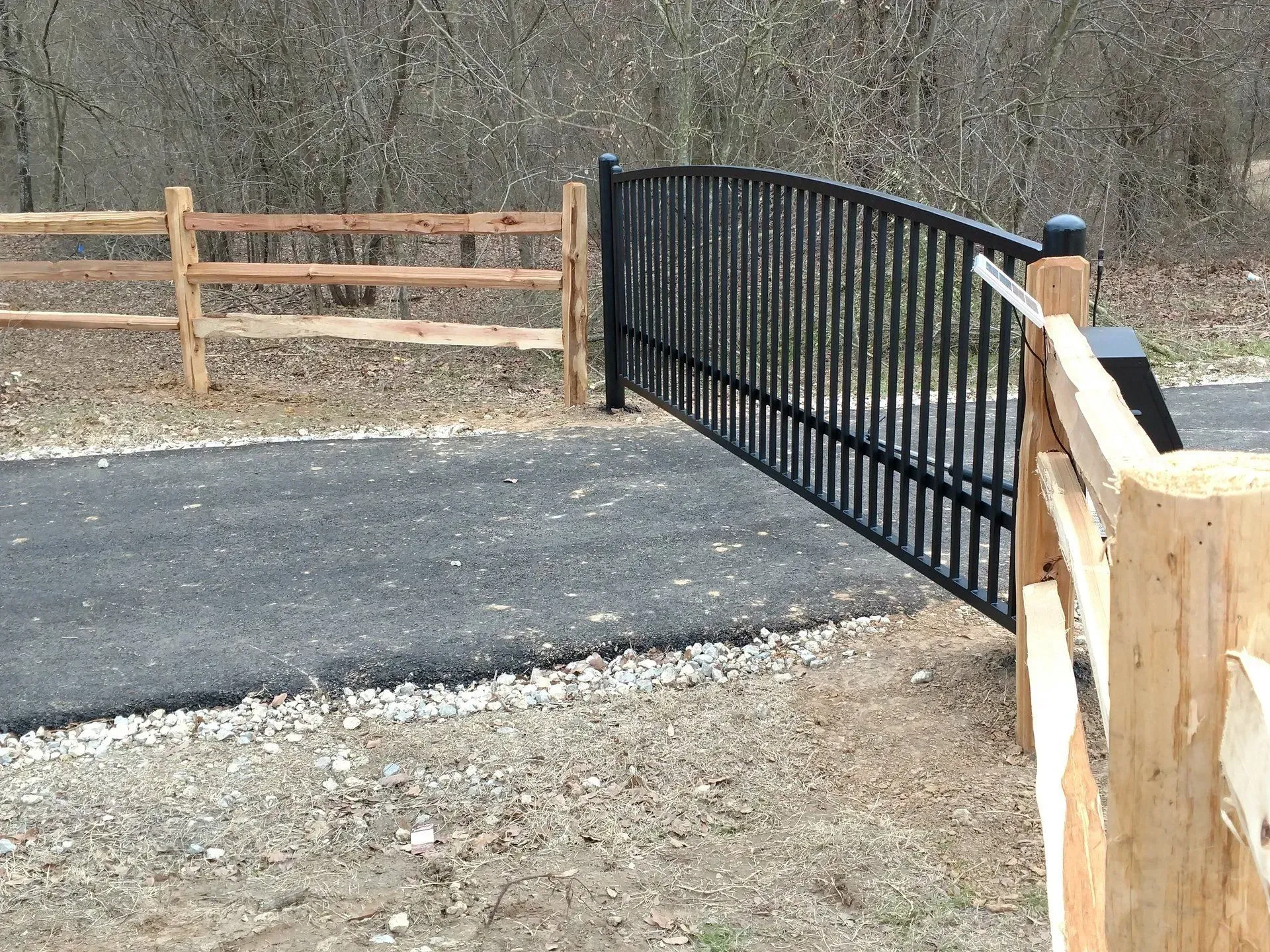 Black gate between wood fences on a paved path in a wooded area.