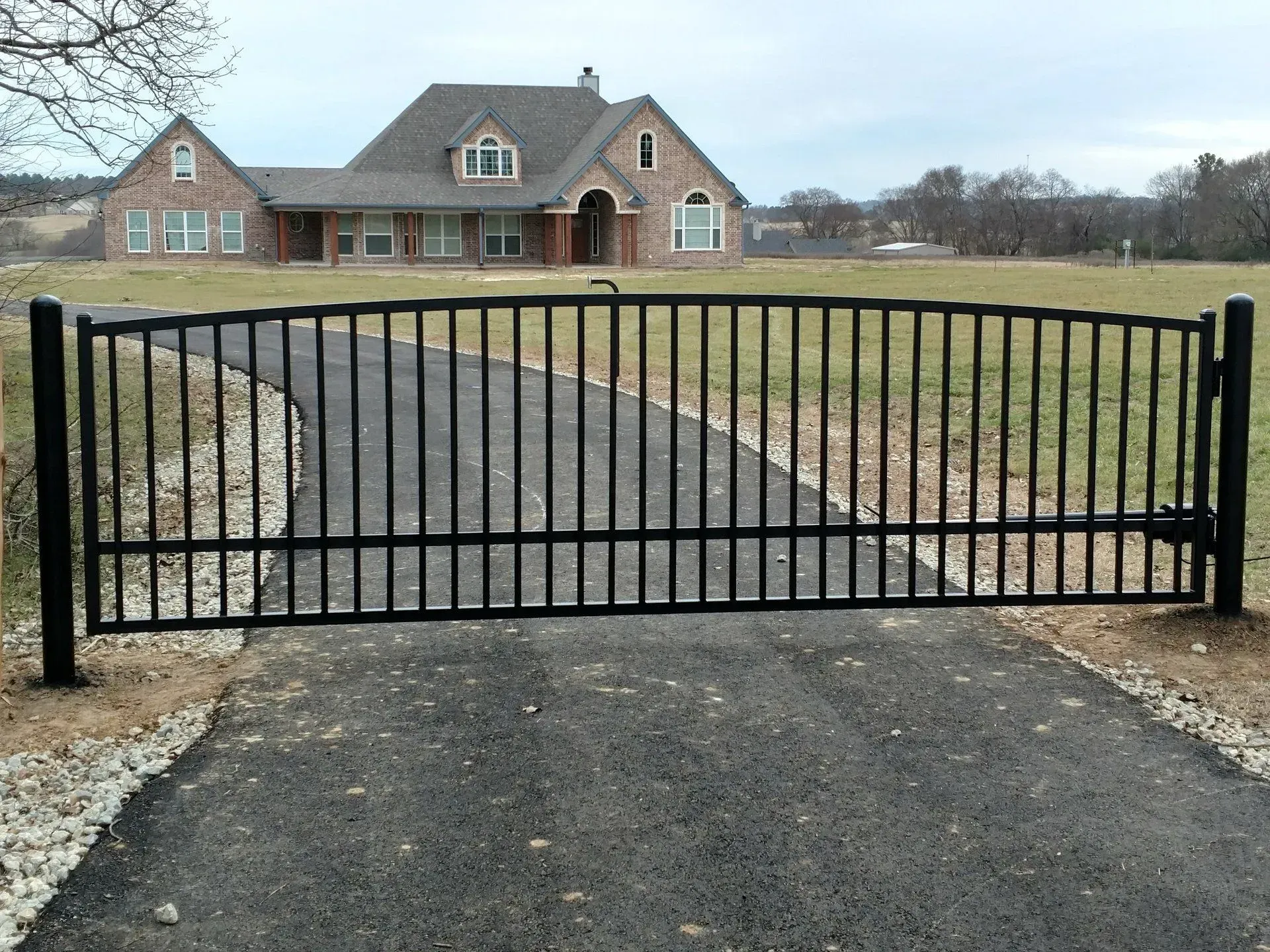 Black iron gate across a paved driveway leading to a brick house.