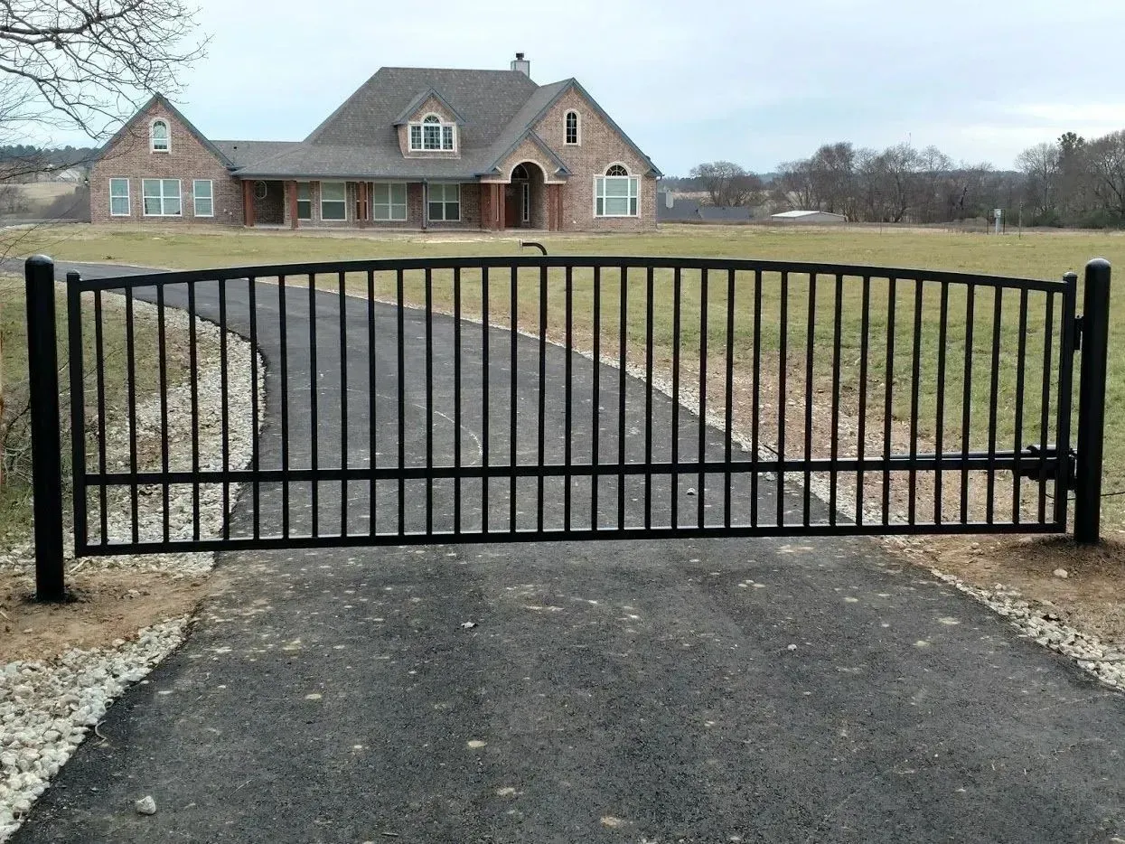 Black metal gate across a paved driveway, leading to a brick house in a rural setting.