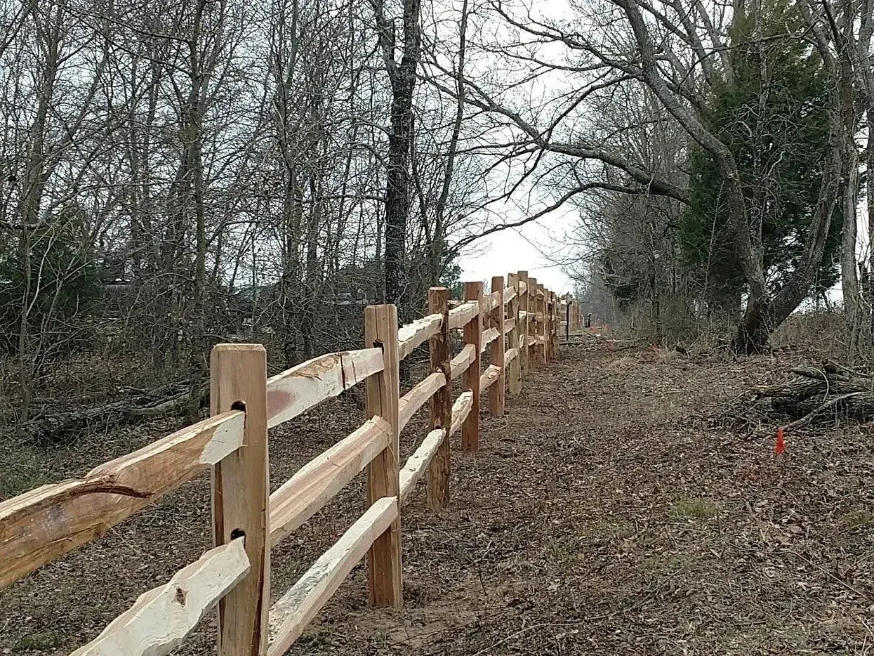 Wooden split-rail fence along a path in a wooded area, on a cloudy day.