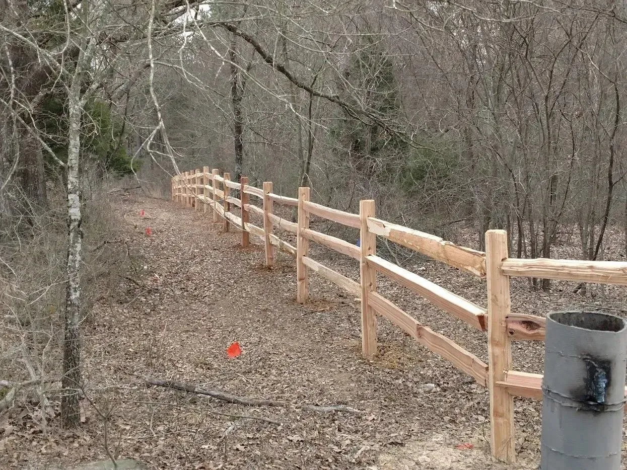 Wooden split-rail fence in a wooded area; leaves on the ground and bare trees.