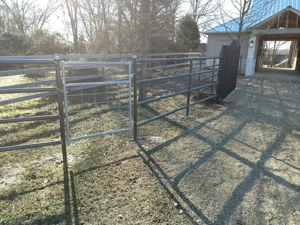 Metal livestock gate in a grassy yard, with a building in the background and a carport to the right.