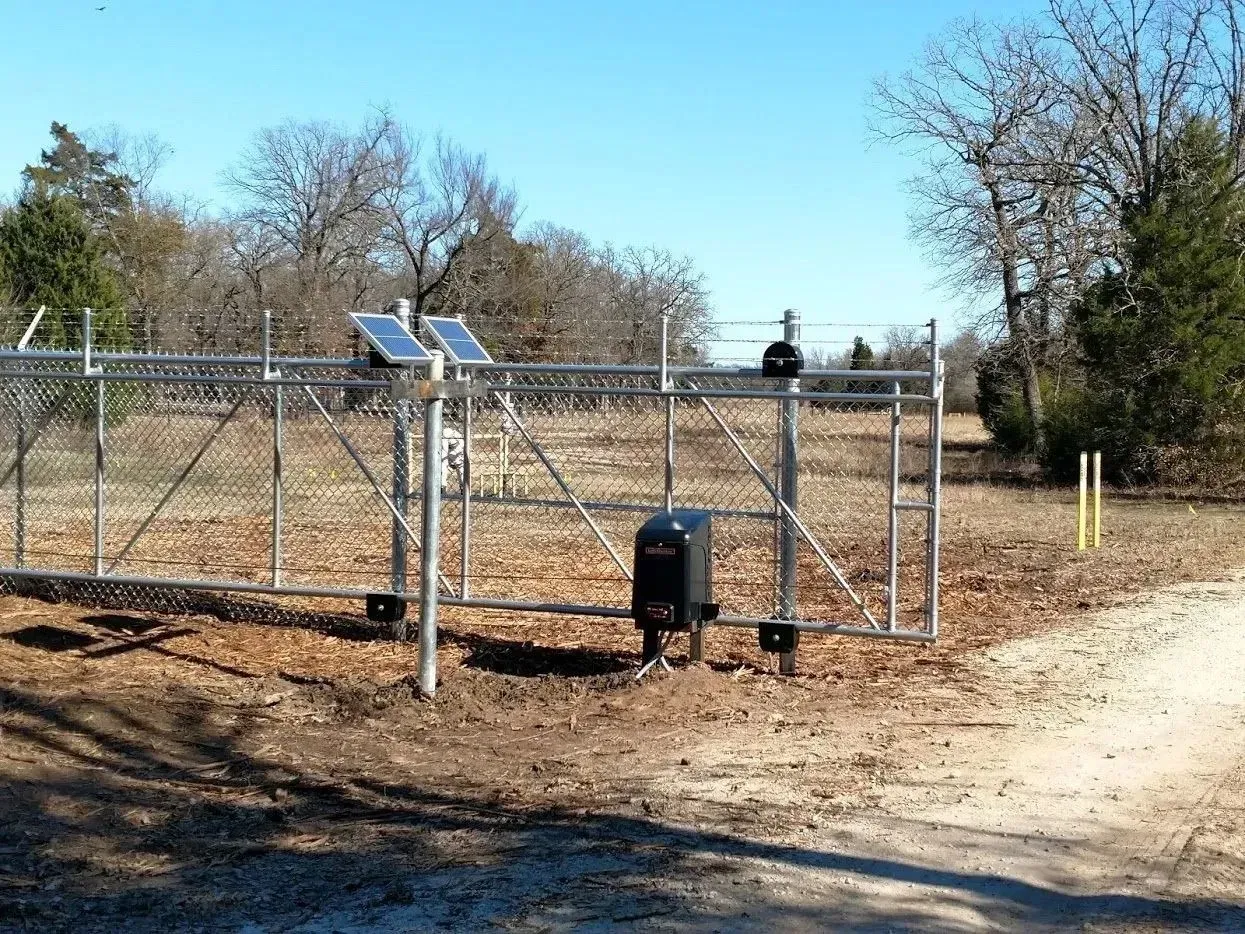 Solar-powered automatic gate in a field. Chain link fence, brown ground, blue sky, and trees.