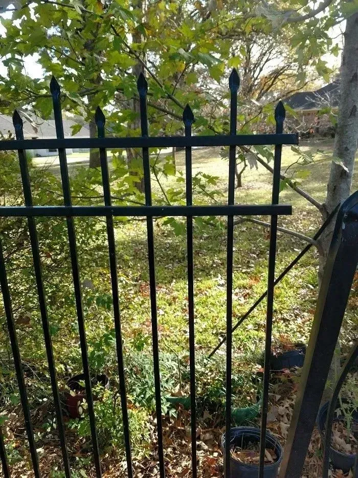 Black metal fence with pointed tops, partially open, with greenery in the background.