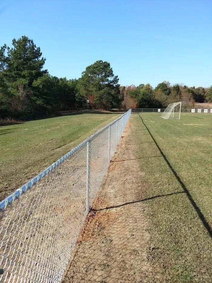 Chain link fence bordering a grassy field with a soccer goal on a sunny day.