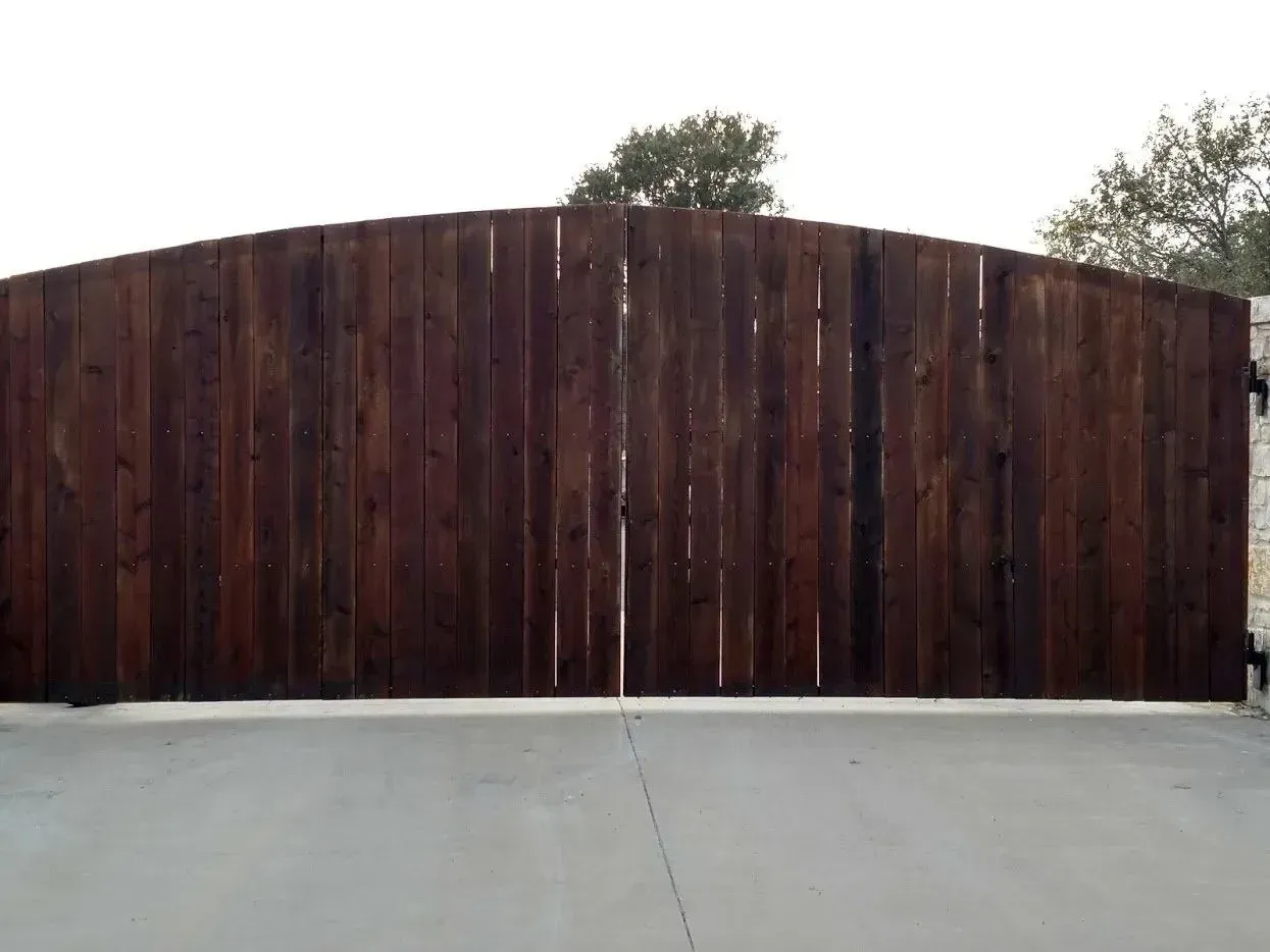 Wooden, arched gate in a concrete driveway; trees and sky in the background.