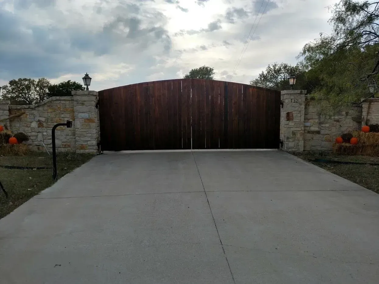 Wooden gate with stone pillars and a concrete driveway under a cloudy sky.