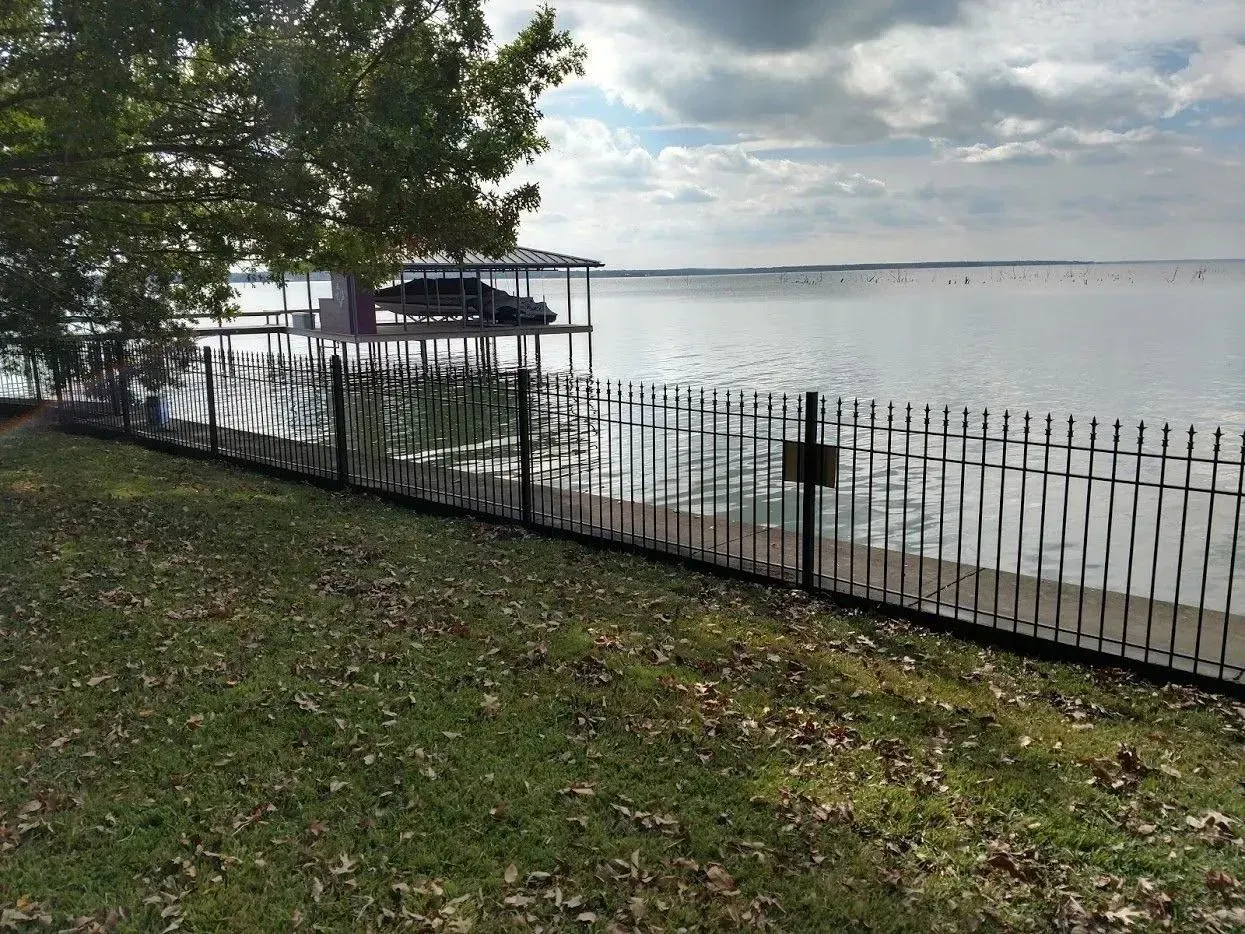 Black fence borders a grassy area leading to a lake with a dock and boat. Cloudy sky.