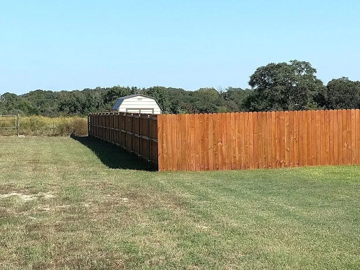 Wooden fence surrounds a grassy yard with a building and trees in the distance under a blue sky.