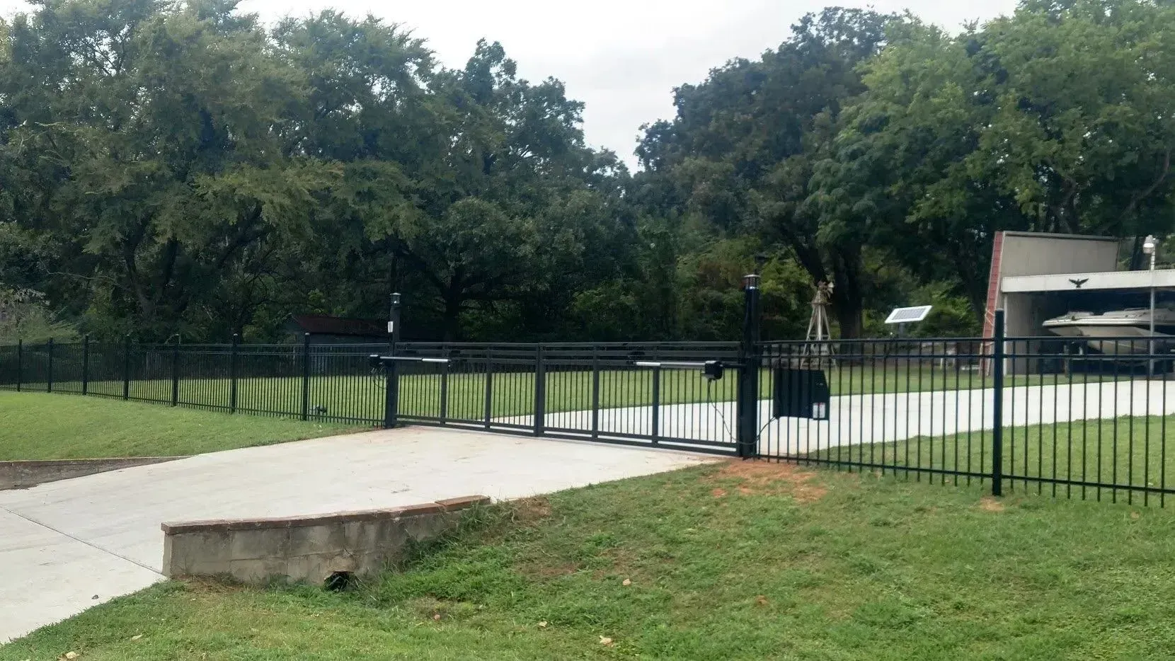 Black gated entrance to a property, concrete driveway, green grass, trees in the background.