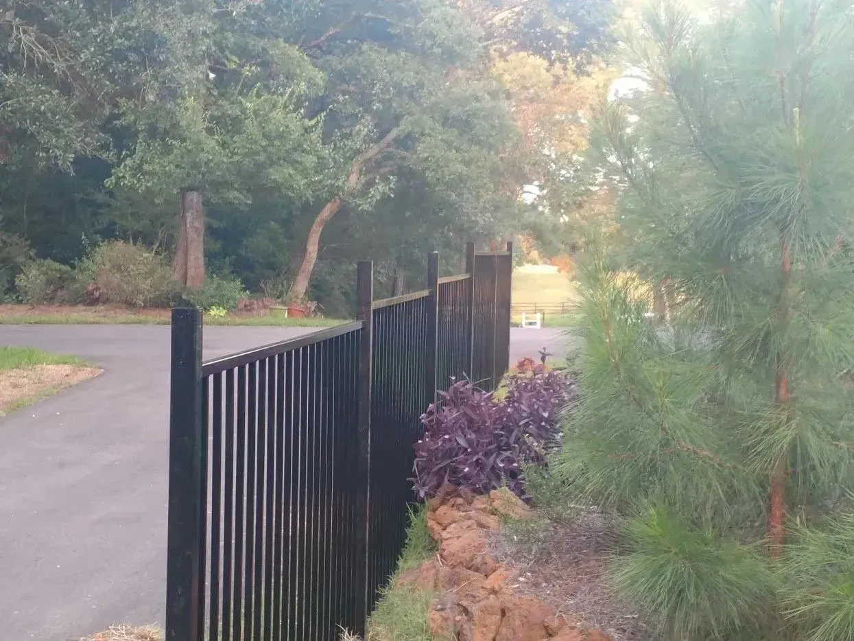 Black fence lines a paved path, beside a rock garden with purple plants and tall trees in the background.