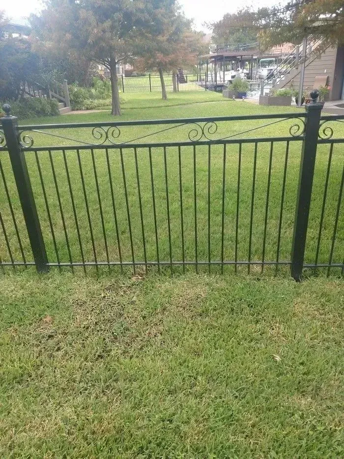 Black wrought-iron fence in a grassy yard, angled slightly, with trees and a house in the background.