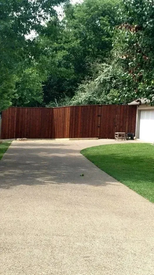 Brown wooden fence in a yard, with a driveway and green grass. Tall trees in the background.