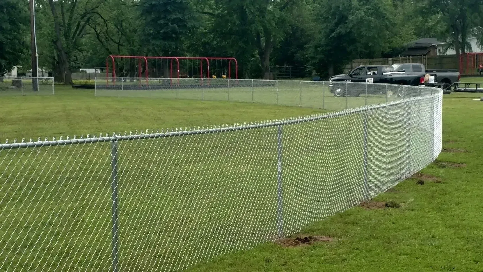 Chain-link fence surrounding a grassy area with a playground and vehicles in the background.