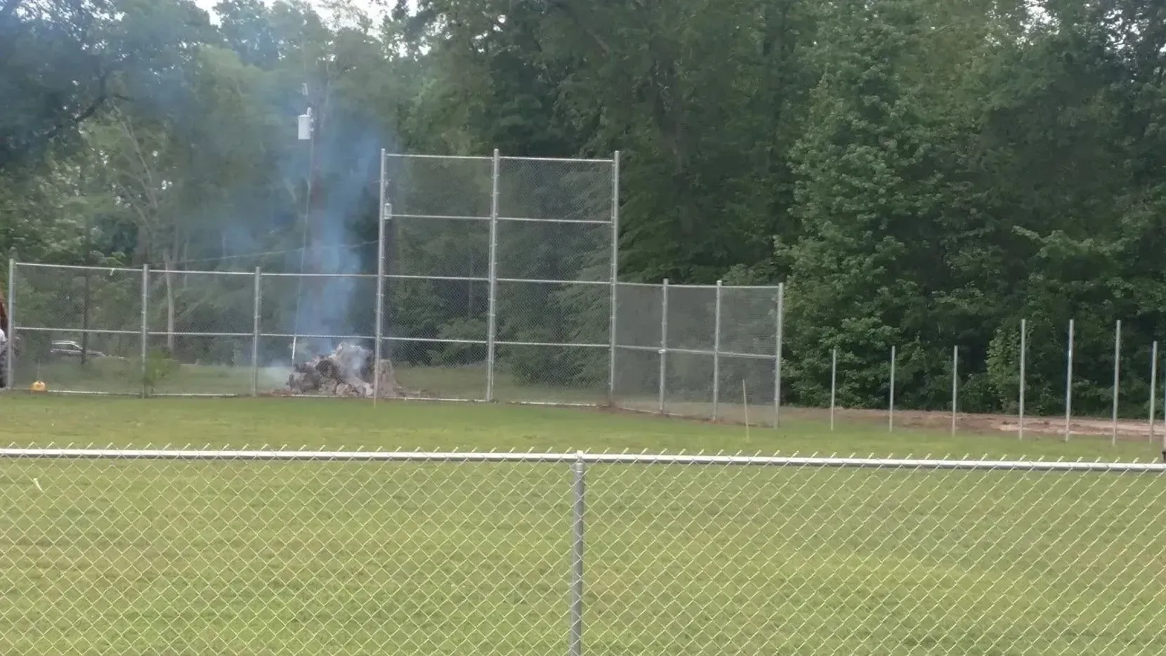 Smoke billows from a fire inside a chain-link fence, set in a grassy field, near trees.