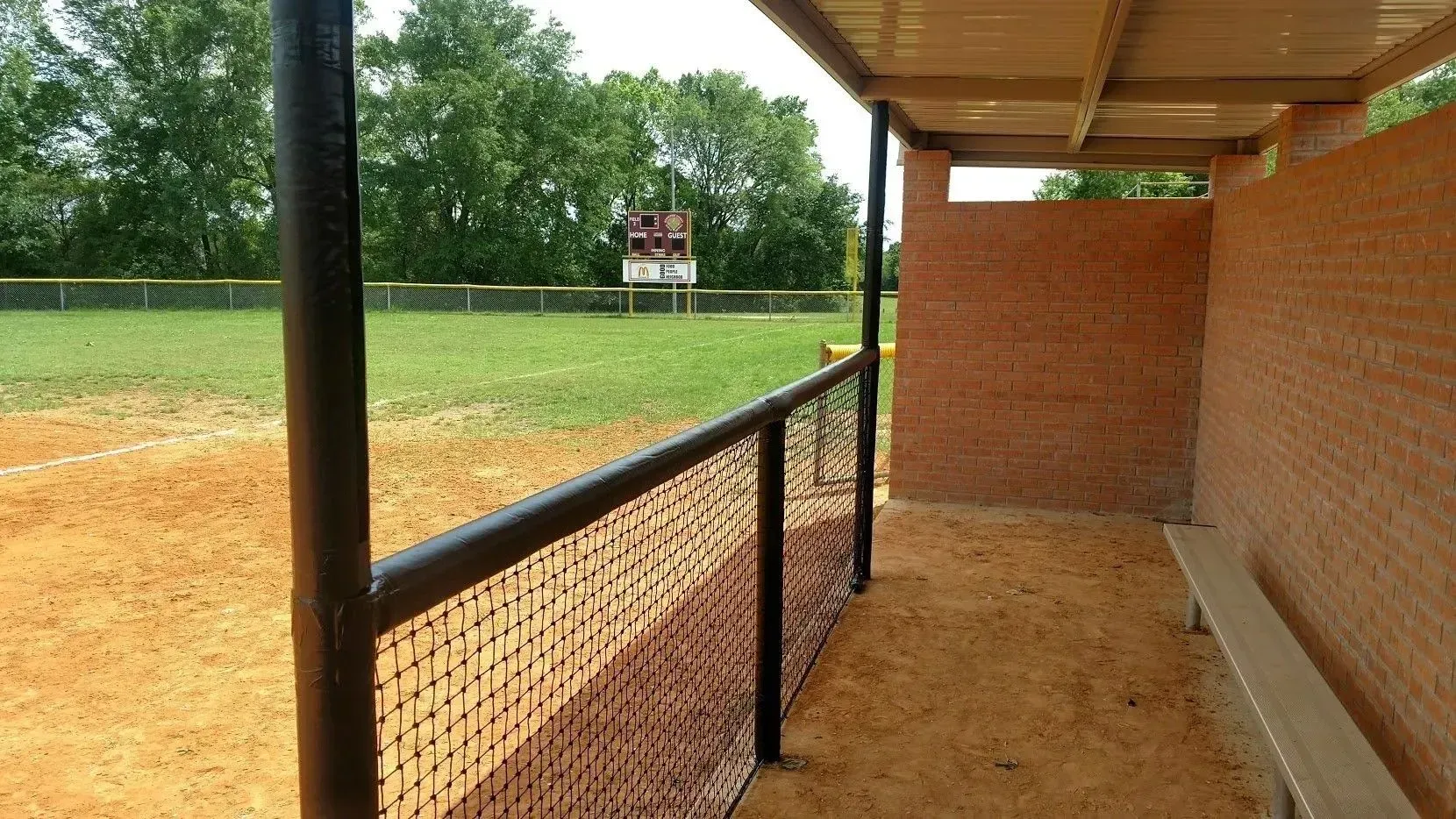 Baseball dugout with bench seating, view of baseball field, scoreboard, and trees in the background.