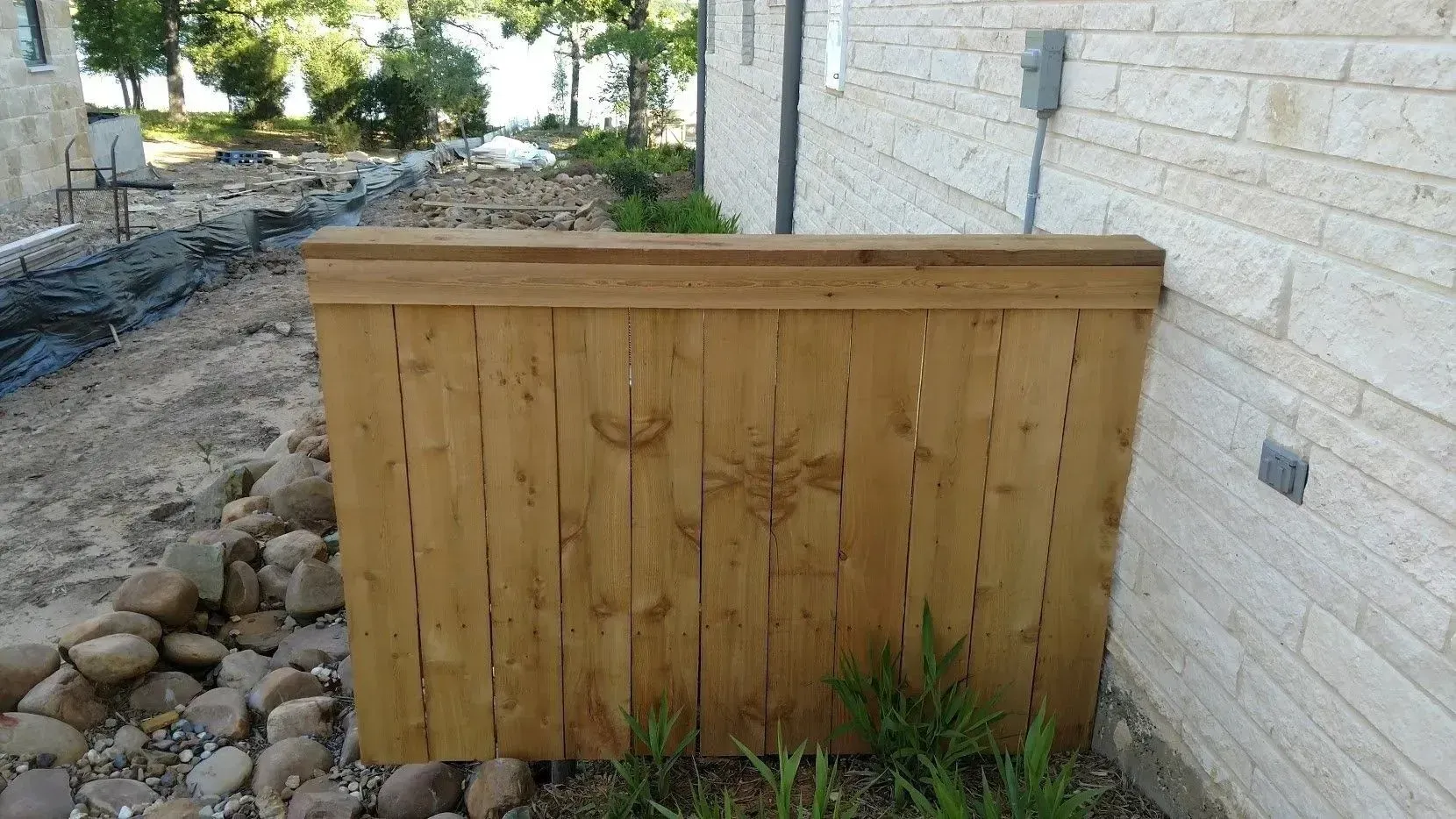 Wooden barrier next to a textured building. Rocks and vegetation are in front of it, trees in the background.