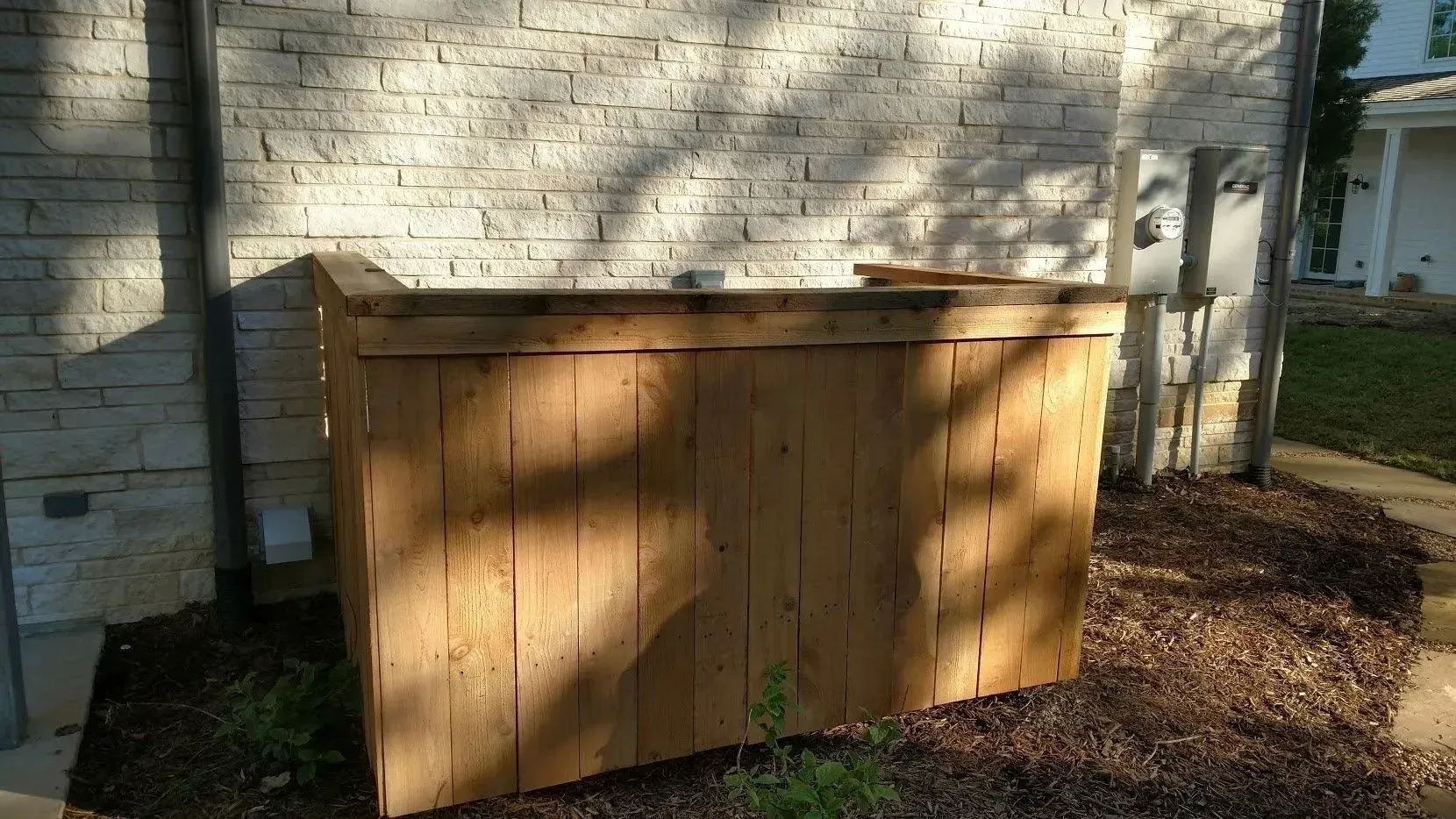 Wooden enclosure in front of a textured stone wall. Brown mulch and plants surround the base.