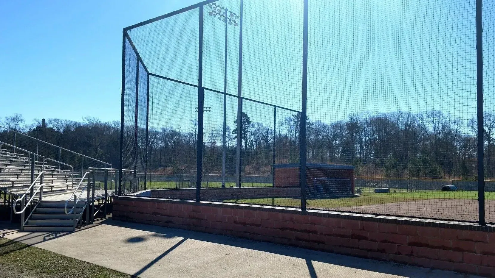 Baseball field with tall backstop, bleachers, and trees under a blue sky.