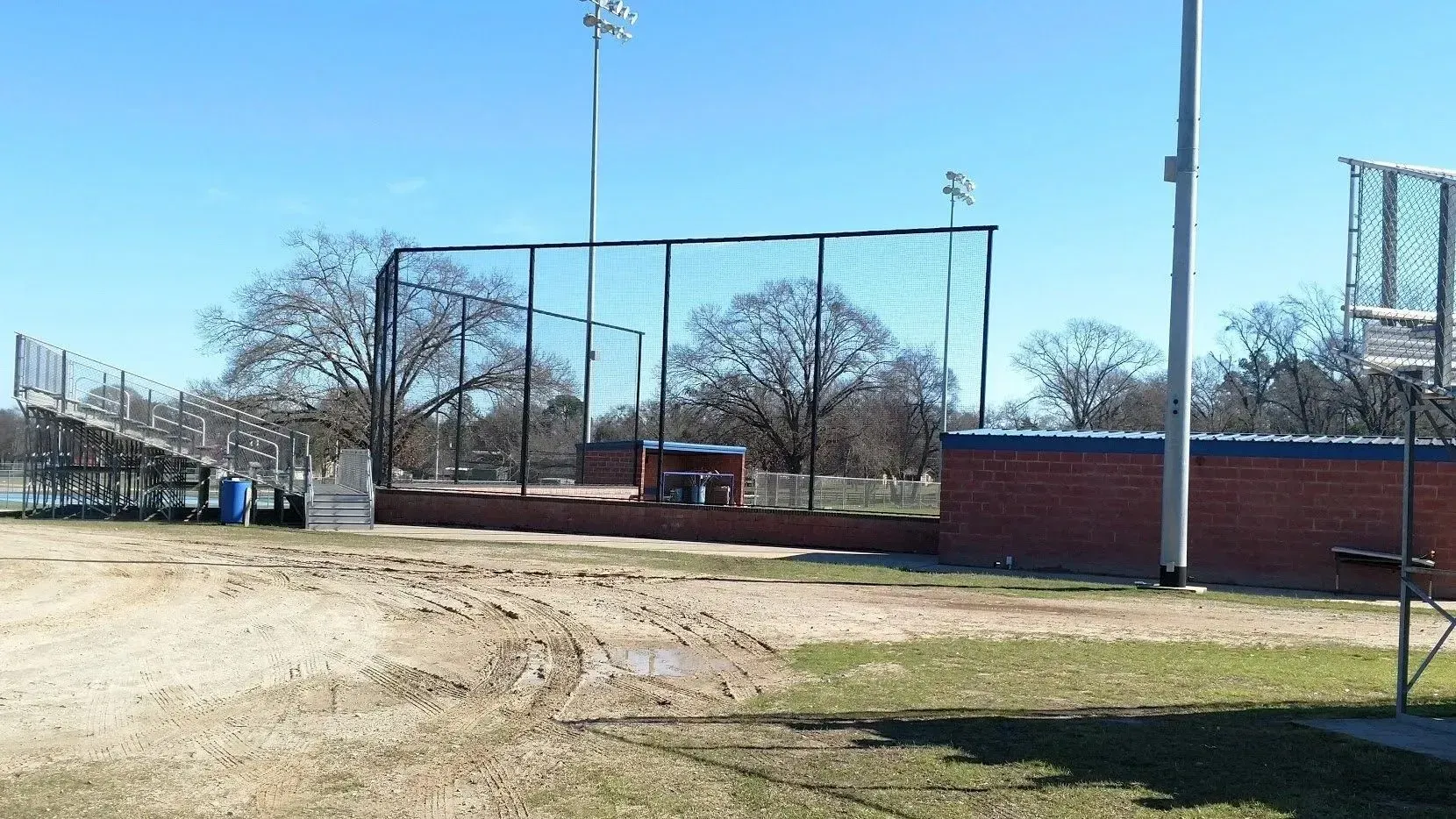 Baseball field, with batting cage, brick wall, bleachers, and tall light poles under a clear blue sky.