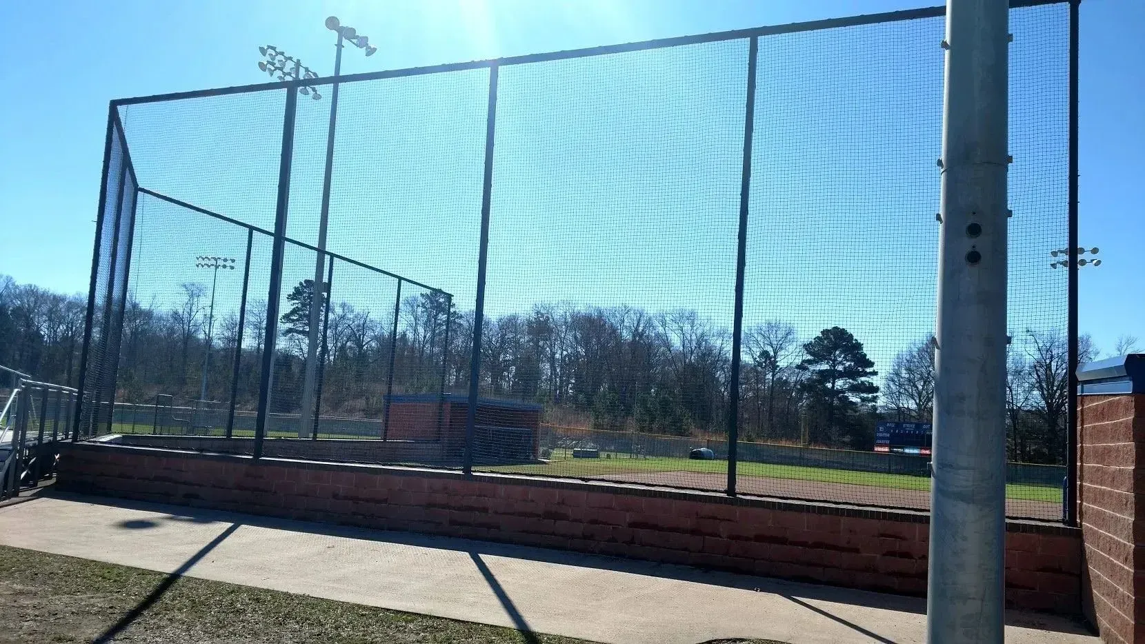 A baseball backstop with black netting, brick wall, and tall light poles against a blue sky.