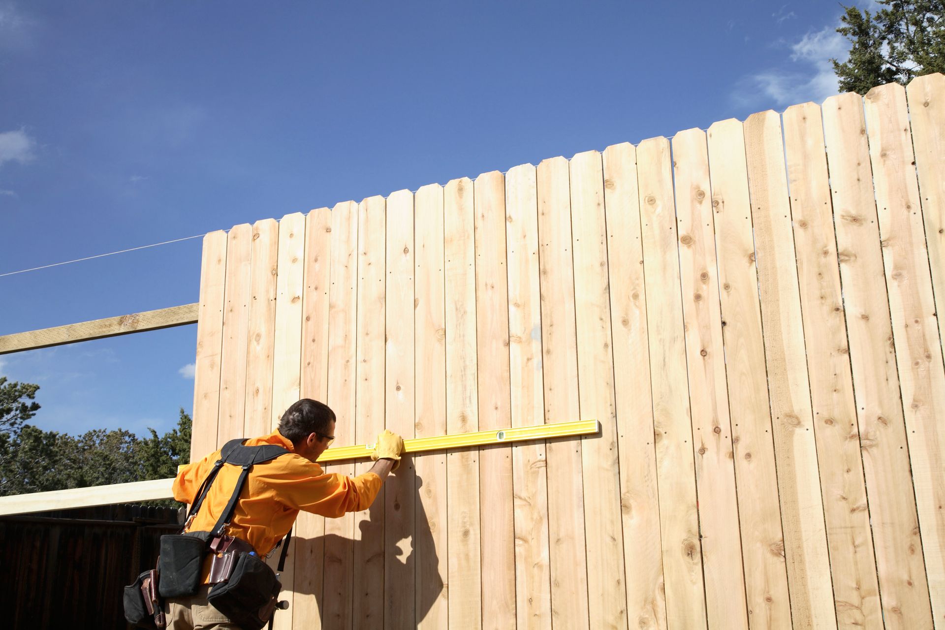Man using a level on a wooden fence outdoors. Bright blue sky.