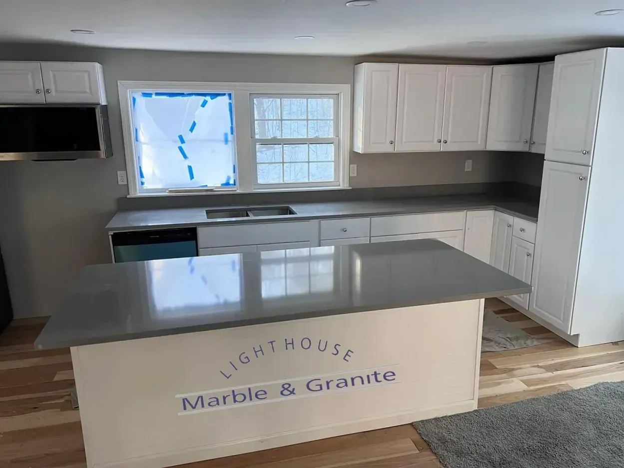 Kitchen with white cabinets, gray countertops, island, and window. "Lighthouse Marble & Granite" logo visible.