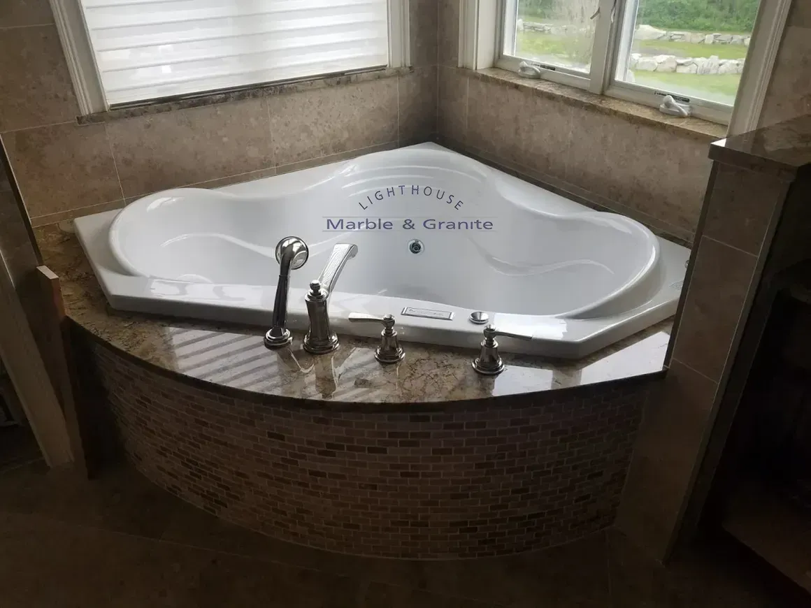 Corner bathtub with mosaic tile surround, granite countertop, and window in background.