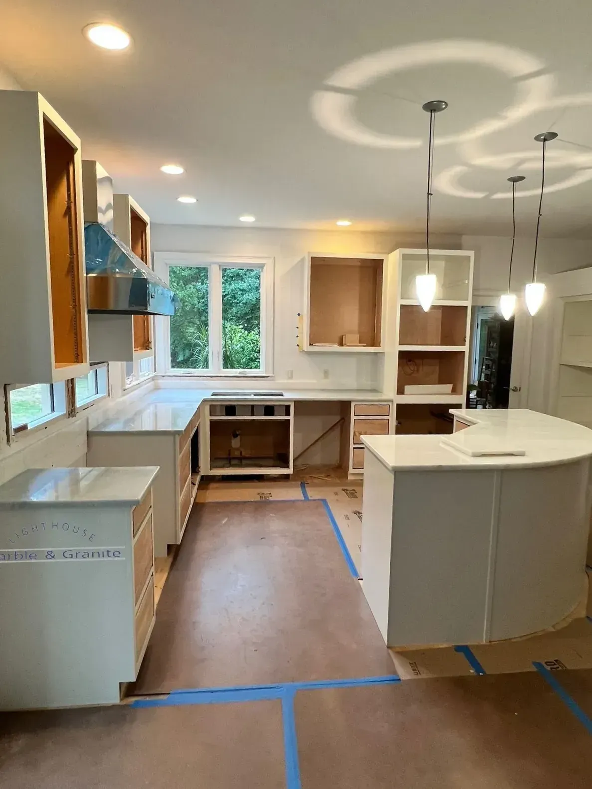 Kitchen renovation in progress: white cabinets, countertops, and island; brown floors; window with green view.