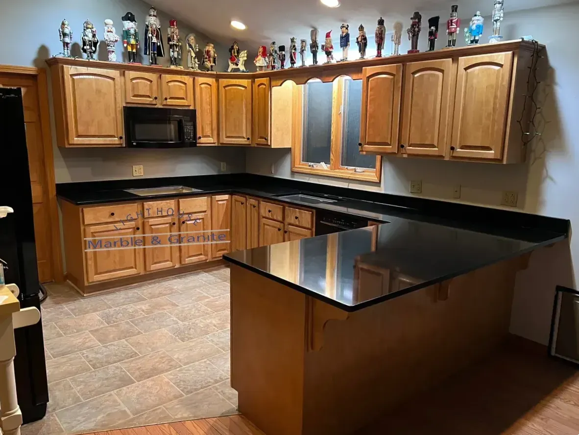 A kitchen with light brown cabinets, black countertops, and a center island, featuring a microwave and figurines on top of cabinets.